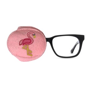 May include: A pink eye patch with a flamingo design attached to black spectacles. The flamingo is embroidered in pink and yellow thread. The spectacles have a rectangular frame.