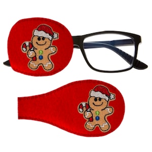 May include: Two red felt eye patches featuring a gingerbread man design. The gingerbread man is wearing a red and white Santa hat and holding a candy cane. The eye patches are designed to be worn over glasses.