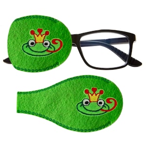 Może przedstawiać: Para oczu z zielonego filcu z haftowanym żabą w koronie na każdym z nich. Plastry są zaprojektowane do zakrywania jednego oka naraz.