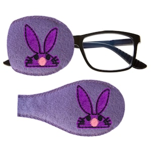 Peut inclure: Deux patchs oculaires en feutre violet avec un motif de lapin rose brodé. Le lapin a un nez rose et des moustaches noires.