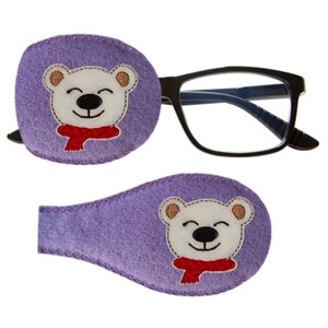 Könnte beinhalten: Zwei violette Filz-Augenklappen mit einem weißen Eisbären, der einen roten Schal trägt, der auf jedem Patch gestickt ist. Die Augenklappen sind so konzipiert, dass sie über den Augen getragen werden, um bei der Behandlung von Amblyopie zu helfen.