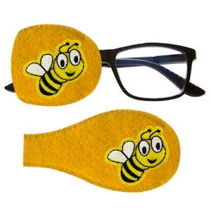 May include: Two yellow felt eye patches with a smiling bee embroidered on each. The bee is black and yellow with white wings.