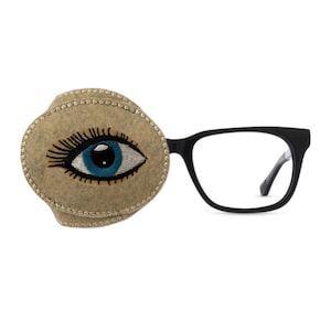 May include: An eye patch with an embroidered eye design attached to a pair of black spectacles. The eye patch is beige with a blue eye and black eyelashes. The spectacles have a rectangular frame.
