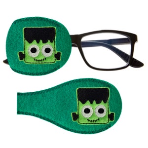 Könnte beinhalten: Zwei grüne Filz-Augenklappen mit einem aufgestickten Cartoon-Frankenstein-Monster-Design. Die Augenklappen sind so konzipiert, dass sie über Brillen getragen werden können.