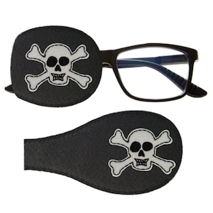 Könnte beinhalten: Zwei schwarze Augenklappen mit einem weißen Totenkopf- und Knochenkreuz-Design. Die Augenklappen sind aus weichem Stoff gefertigt und zum Tragen über dem Auge konzipiert.