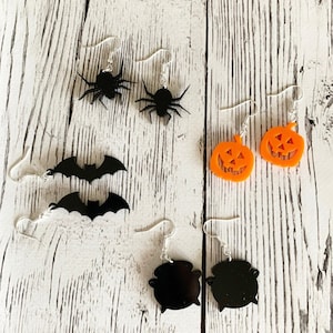 May include: A set of Halloween-themed earrings featuring black bats, spiders, cauldrons, and orange pumpkins. The earrings are made of acrylic and have silver hooks.