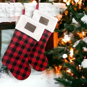 May include: Two Christmas stockings with red and black buffalo plaid patterns. Each stocking has a white faux fur cuff and a brown leatherette tag with a name on it. One says "Travis" and the other says "Dezarne".