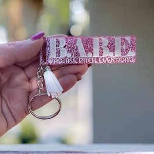 May include: Pink glitter keychain with a white tassel and the text "BABE BAD ASS. BITCH. EVERYDAY."