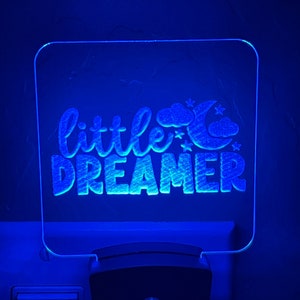 May include: A blue night light with the words "little dreamer" in a script font. The design includes a crescent moon, clouds, and stars. The night light is plugged into a wall outlet.