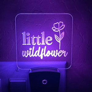 May include: A square, clear acrylic night light with the words "little wildflower" and a floral design, illuminated with a purple glow. The night light plugs into a wall outlet.