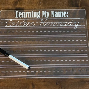 Puede incluir: Una pizarra de borrado en seco de plástico transparente con el texto "Learning My Name:" y el nombre "Caiden Hernandez" escrito en cursiva. Un marcador de borrado en seco blanco está sobre la pizarra.
