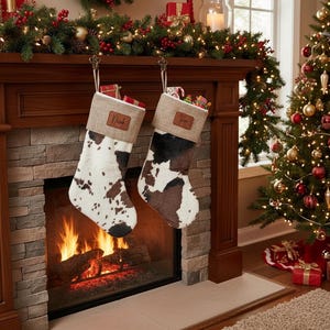 May include: Two Christmas stockings with a cowhide pattern hang from a wooden mantel. One stocking is white and brown, the other is black and brown. The stockings are personalized with names on leather tags. A Christmas tree and fireplace are in the background.