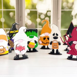 May include: Seven Halloween-themed gnomes made of wood. The gnomes are painted in various colors, including orange, green, white, red, and black. Each gnome has a different Halloween-themed accessory, such as a pumpkin, a candy corn, a candy cane, a ghost, a devil, and a witch's hat.