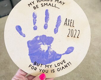 May include: A round wooden plaque with a purple handprint and the text "MY HANDS MAY BE SMALL... AXEL 2022 BUT MY LOVE FOR YOU IS GIANT!"