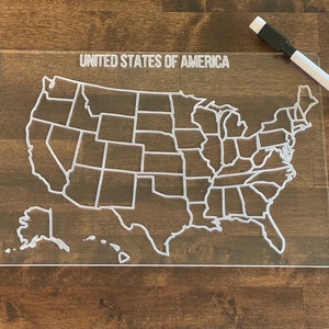 May include: A clear acrylic dry-erase map of the United States with the states outlined in white. The map includes Alaska and Hawaii. The text "UNITED STATES OF AMERICA" is printed at the top of the map.