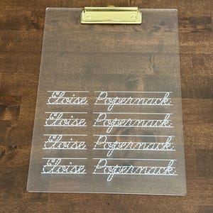 May include: A clear acrylic clipboard with a gold metal clip. The name "Elise Peppermack" is written in white cursive on the clipboard.