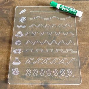May include: A clear plastic dry-erase board with white lines and various images, including a butterfly, a worm, a frog, a pumpkin, a crab, a sailboat, and a bee. The board is designed for tracing and practicing fine motor skills. The board is accompanied by a green dry-erase marker with the text "Expo low odor dry erase chisel" on the side.