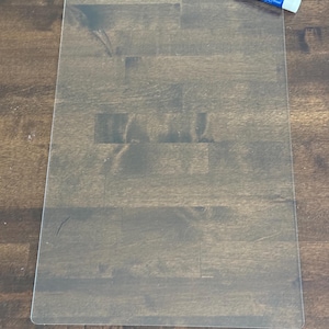 May include: A clear, rectangular dry-erase board with rounded corners. A blue and white Expo low odor chisel tip marker is in the upper right corner. The board is on a dark brown wooden surface.
