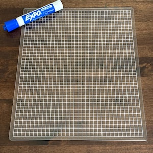 May include: A clear, grid-marked cutting mat with a blue and white Expo dry erase marker. The marker has the words "low odor" and "chisel" printed on it. The mat is rectangular and has a grid pattern.