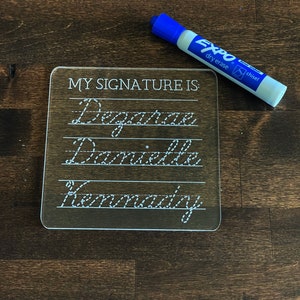 May include: A clear plastic dry erase board with the text "MY SIGNATURE IS:" at the top. The board has three lines with dotted lines for practice writing the words "Dagmar", "Danielle", and "Kennedy". A blue dry erase marker with the brand name "Expo" is next to the board.