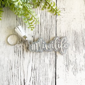May include: A silver glitter keychain with the text "#MimiLife" in white script. The keychain has a white tassel and a silver keyring. The background is a white wooden surface with green foliage.