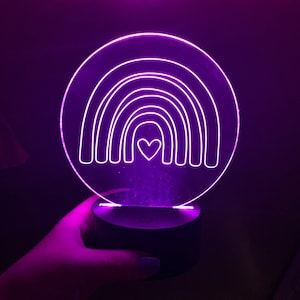 May include: A glowing purple LED rainbow night light with a heart in the center. The rainbow design is etched into a clear acrylic circle, mounted on a black base. The light emits a soft, ambient glow.