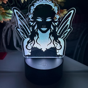 May include: A white fairy silhouette night light with a crown and wings. The light is on a black base with a power button.