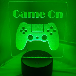 May include: A green-lit acrylic gaming sign with the words "Game On" above a controller graphic. The sign is mounted on a black circular base with a power button symbol. The overall design is illuminated with a bright green glow.
