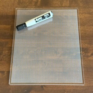May include: A clear plastic grid with a white and black Expo dry-erase marker. The grid has a square pattern and is on a wooden surface. The marker is white with black text and a black tip.