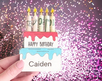 May include: A wooden birthday cake with pink and blue frosting and candles made of rolled up dollar bills. The cake says "Happy Birthday Caiden" on it.