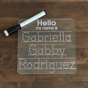 May include: A clear plastic dry-erase board with dotted lines for tracing letters. The board says "Hello my name is" and has the name "Gabriella Gabby Rodriguez" written on it. A black and white dry-erase marker is next to the board.
