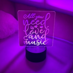 May include: A white acrylic night light with the text "All you need is love and music" in a cursive font. The light is on and illuminating the text in a purple hue.