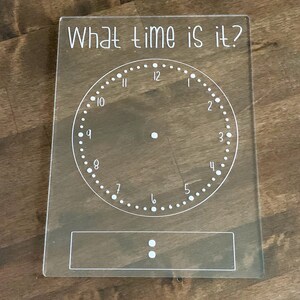 May include: A white acrylic clock face with black numbers and a white rectangle with two dots below. The clock face says "What time is it?" at the top.