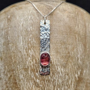 Puede incluir: Un colgante rectangular de plata con un acabado martillado y texturizado que presenta una piedra preciosa rosa de forma ovalada engastada cerca de la base. El colgante cuelga de una fina cadena de plata, creando una pieza de joyer&iacute;a vertical.