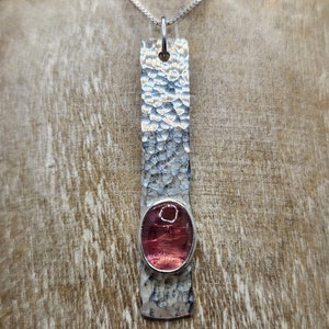 Puede incluir: Un colgante rectangular de plata con una superficie martillada y texturizada, que presenta una piedra preciosa rosa transl&uacute;cida de forma ovalada engastada en un bisel liso. El colgante cuelga de una fina cadena de plata, creando una pieza vertical.