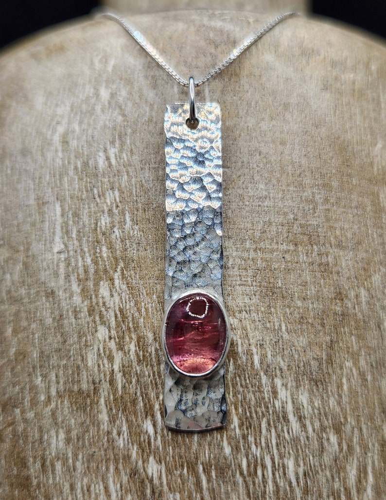 Puede incluir: Un colgante rectangular de plata con una superficie martillada y texturizada, que presenta una piedra preciosa rosa transl&uacute;cida de forma ovalada engastada en un bisel liso. El colgante cuelga de una fina cadena de plata, creando una pieza vertical.