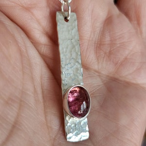 Puede incluir: Un colgante de plata con una barra larga y rectangular de acabado martillado y texturizado. Una piedra preciosa ovalada de color rosa intenso est&aacute; engastada cerca de la parte inferior de la barra de plata. El colgante cuelga de una cadena fina.