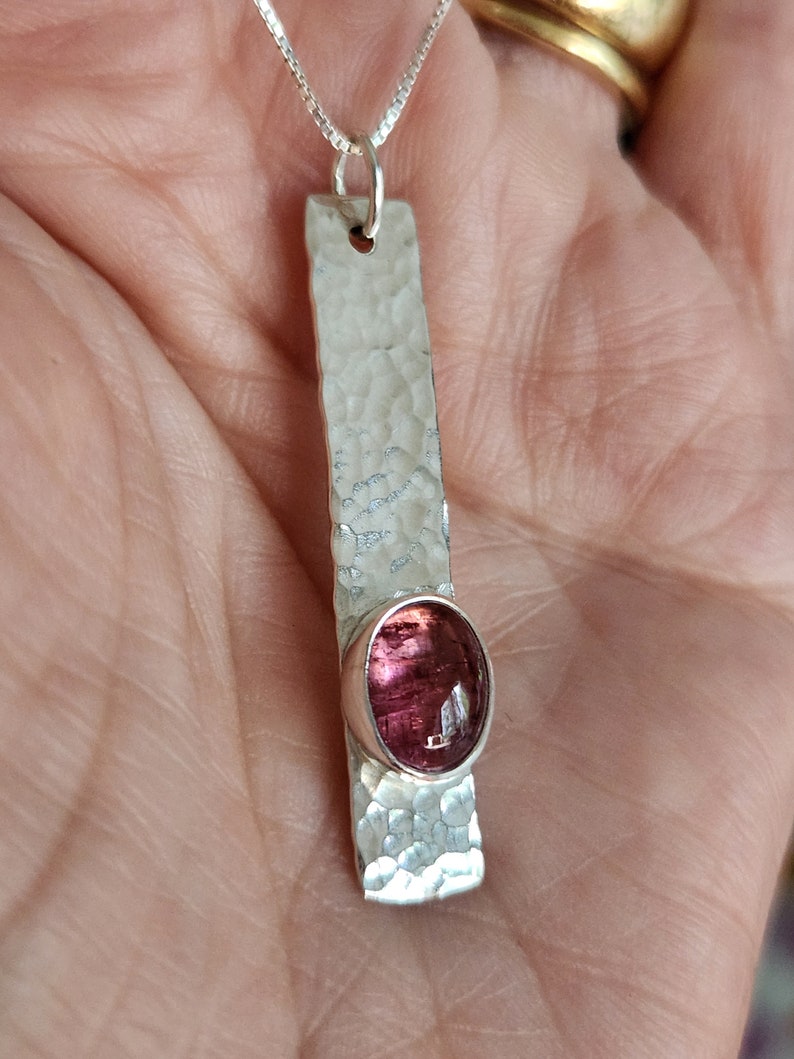 Puede incluir: Un colgante de plata con una barra larga y rectangular de acabado martillado y texturizado. Una piedra preciosa ovalada de color rosa intenso est&aacute; engastada cerca de la parte inferior de la barra de plata. El colgante cuelga de una cadena fina.