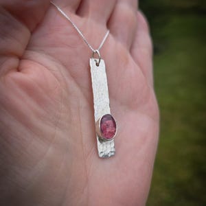 Puede incluir: Un collar con colgante de plata que presenta una barra met&aacute;lica rectangular larga con una superficie martillada y texturizada. Una piedra preciosa de color rosa intenso y forma ovalada est&aacute; engastada cerca de la base de la barra, que cuelga de una fina cadena de plata.