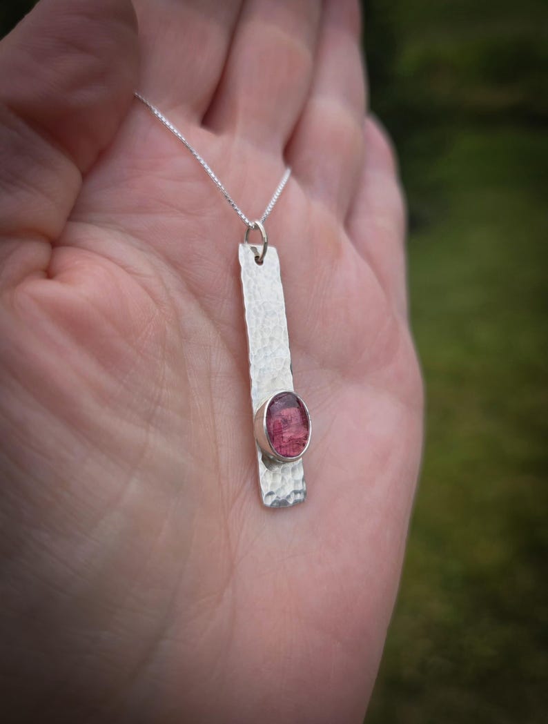 Puede incluir: Un collar con colgante de plata que presenta una barra met&aacute;lica rectangular larga con una superficie martillada y texturizada. Una piedra preciosa de color rosa intenso y forma ovalada est&aacute; engastada cerca de la base de la barra, que cuelga de una fina cadena de plata.