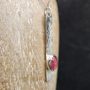 Puede incluir: Un collar con colgante de plata que presenta una barra met&aacute;lica larga y rectangular con una superficie martillada y texturizada. Una piedra preciosa redonda de color rosa intenso est&aacute; engastada en un bisel de plata liso en la parte inferior.