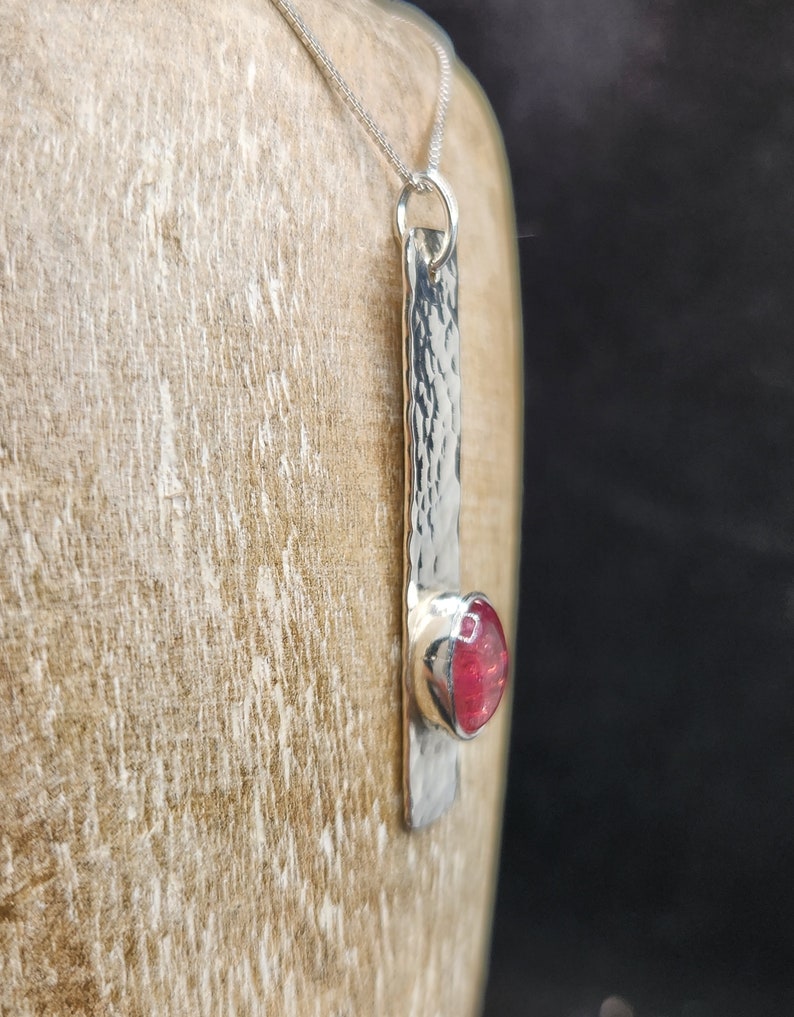 Puede incluir: Un collar con colgante de plata que presenta una barra met&aacute;lica larga y rectangular con una superficie martillada y texturizada. Una piedra preciosa redonda de color rosa intenso est&aacute; engastada en un bisel de plata liso en la parte inferior.