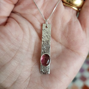 Puede incluir: Un colgante rectangular de plata con textura martillada y una piedra preciosa rosa de forma ovalada engastada cerca de la base. El colgante cuelga de una fina cadena de plata, creando un dise&ntilde;o vertical que resalta el contraste del metal.