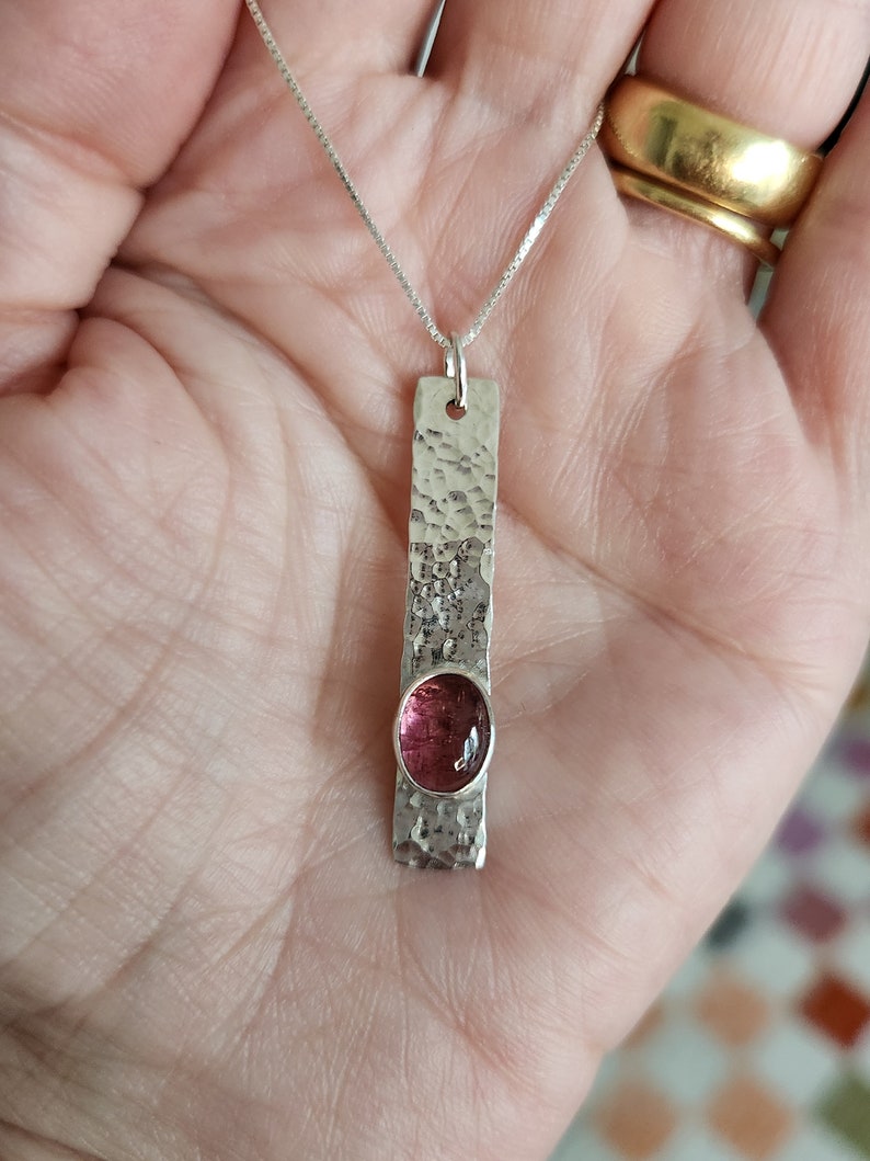 Puede incluir: Un colgante rectangular de plata con textura martillada y una piedra preciosa rosa de forma ovalada engastada cerca de la base. El colgante cuelga de una fina cadena de plata, creando un dise&ntilde;o vertical que resalta el contraste del metal.