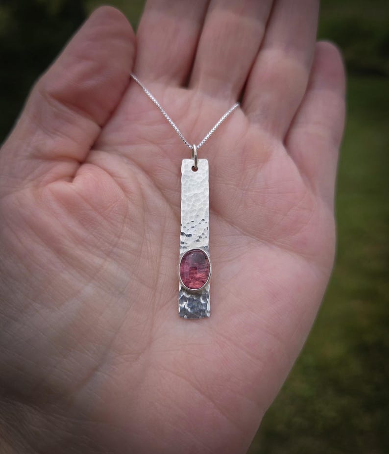 Puede incluir: Un colgante rectangular de plata con una superficie martillada y texturizada que presenta una piedra preciosa rosa de forma ovalada en la mitad inferior. El colgante cuelga de una delicada cadena de plata, creando una pieza de joyer&iacute;a sencilla.