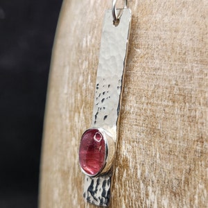 Puede incluir: Un collar con colgante de plata que presenta una barra de metal larga y rectangular con un acabado martillado y texturizado. Una piedra preciosa redonda y rosa transl&uacute;cida est&aacute; engastada cerca de la parte inferior de la barra. El colgante cuelga de una cadena de plata fina.