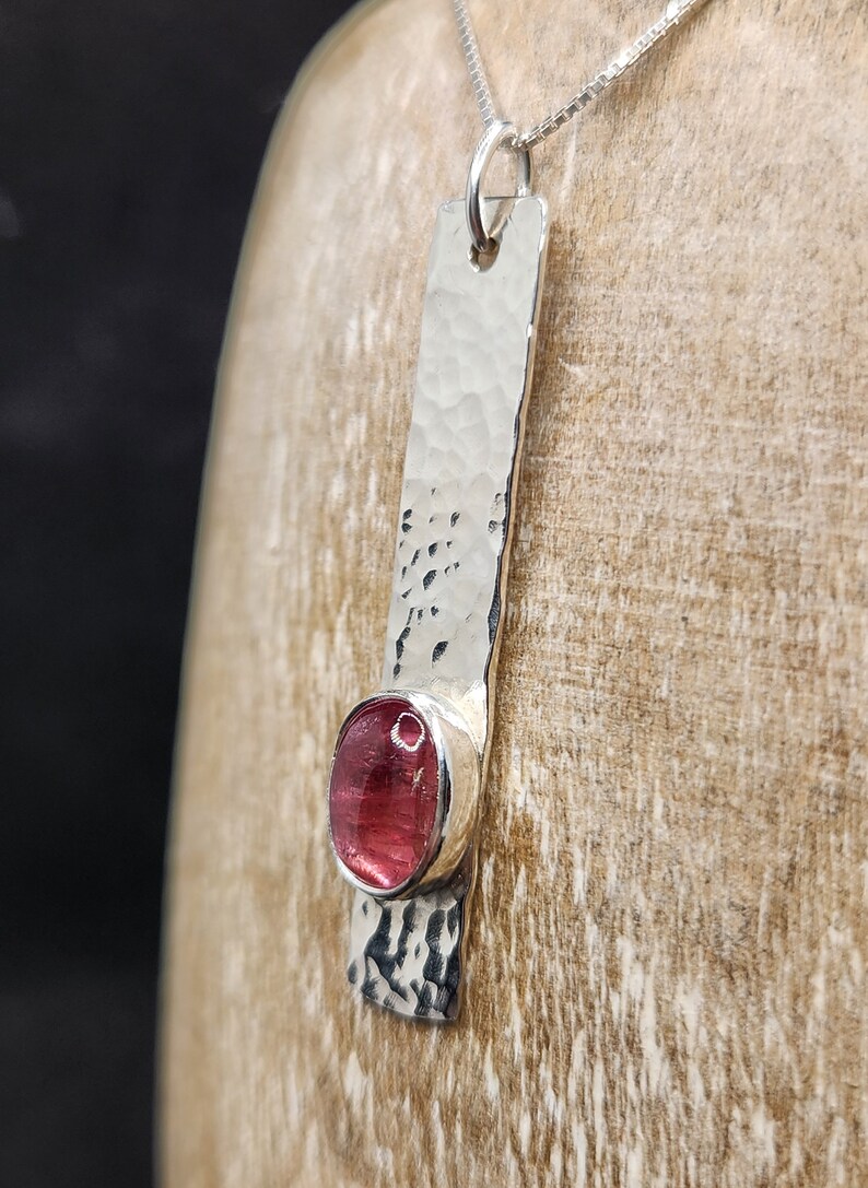 Puede incluir: Un collar con colgante de plata que presenta una barra de metal larga y rectangular con un acabado martillado y texturizado. Una piedra preciosa redonda y rosa transl&uacute;cida est&aacute; engastada cerca de la parte inferior de la barra. El colgante cuelga de una cadena de plata fina.