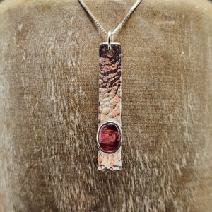 Puede incluir: Un colgante rectangular de metal con textura martillada, p&aacute;tina oscura y acabado pulido. Una piedra preciosa ovalada de color rojo intenso est&aacute; engastada cerca de la parte inferior de la barra vertical. El colgante cuelga de una cadena de plata.
