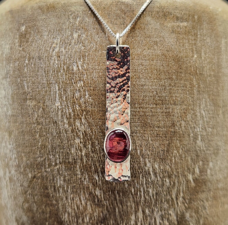Puede incluir: Un colgante rectangular de metal con textura martillada, p&aacute;tina oscura y acabado pulido. Una piedra preciosa ovalada de color rojo intenso est&aacute; engastada cerca de la parte inferior de la barra vertical. El colgante cuelga de una cadena de plata.