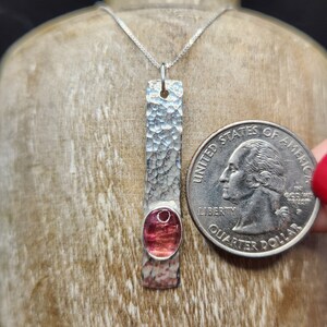Puede incluir: Un collar con colgante de plata que presenta una barra de metal larga, rectangular y martillada con una piedra preciosa rosa ovalada engastada cerca de la base. Una moneda de 25 centavos de EE. UU. se coloca al lado para mostrar la escala.